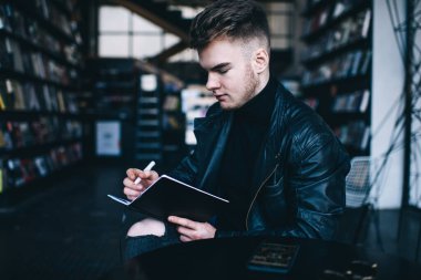 Modern kütüphanede sınavlara hazırlanırken siyah deri ceketli, not defterinden bilgi okuyan konsantre olmamış genç bir erkek öğrencinin yan görüntüsü.