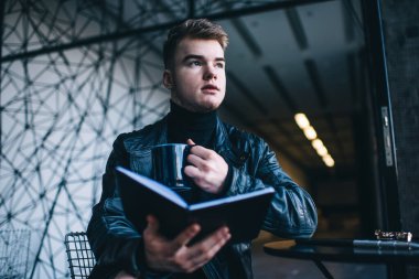 Modern kafede ilginç bir kitap okurken, bir fincan çaydan keyif alan ve dalgın dalgın bakan, modaya uygun giyinmiş genç bir erkek.