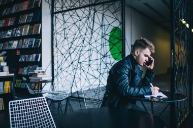 Modern kitapçıda küçük yuvarlak bir masada otururken akıllı telefondan notlar alan modaya uygun deri ceketli, ciddi bir gencin yan görünüşü.