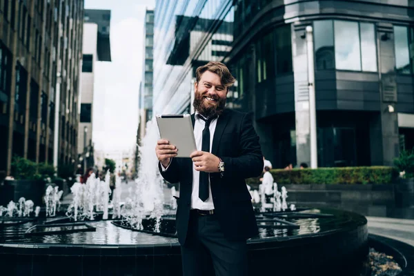 Resmi giysiler içinde neşeli sakallı bir erkek işçi. Gülümsüyor, kameraya bakıyor. Modern sokakta tablet ararken.