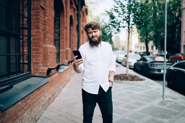 快乐的大胡子男人穿着休闲装站在街上 白天休息时用手机和朋友聊天 — 图库照片