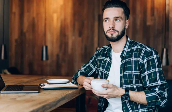 Düşünceli bir erkek blogcu, elinde sıcak kafein içeceği ve düşünceli bakışlarla kafeteryada uzaktan çalışan düşünceli bir hippi ile birlikte teknoloji netbook 'uyla masasında oturuyor.