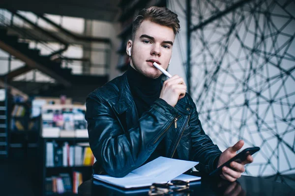 Gerçek kablosuz kulaklıklı genç bir erkek öğrenci kütüphanede açık not defteri ve akıllı telefonla kameraya bakarken dalgın dalgın bakıyor.