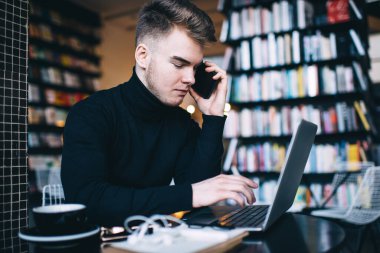 Siyah balıkçı yakalı dalgın bir gencin iş için dizüstü bilgisayar kullanırken ve kütüphaneden uzak bir projede çalışırken telefonla konuşurken yan görüntüsü
