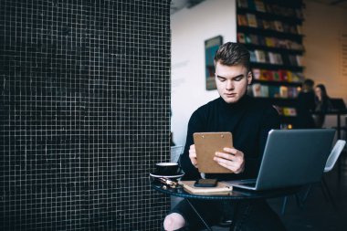 Küçük yuvarlak bir masada dizüstü bilgisayarı ve not panosuyla oturup uzaktan çalışan ve kahve içen serbest çalışan genç bir erkek.