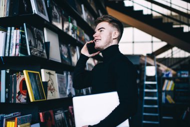 Aşağıdan bakıldığında sıradan kıyafetlerle gülümseyen genç bir öğrencinin cep telefonuyla konuşurken kütüphanenin kütüphanesinde elinde netbook ile dikilip başka yere baktığı görülüyor.