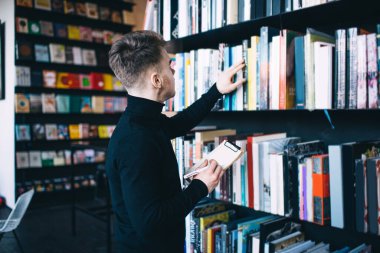 Balıkçı yakalı, modern saç stiliyle kitap arayan, kütüphanede elinde not panosu ve kalemle kitap taşıyan isimsiz bir adamın yan görüntüsü.