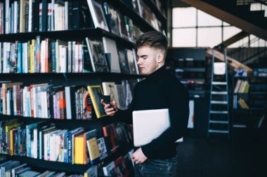 Modern saç stili olan, netbook ile kitaplığın yanında duran, kütüphanenin arkasında cep telefonuyla sohbet eden, ciddi bir erkeğin yan görüntüsü.