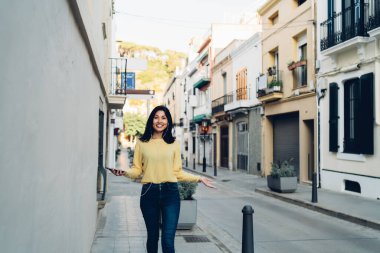 Kulaklıklı neşeli turistin yarım boy portresi turistik şehirde geziyor ve kameraya gülümsüyor, cep telefonu cihazlı neşeli hippi kız boş zamanlarında müzik dinlemek için medya uygulaması kullanıyor.