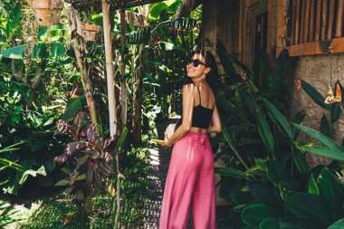 Pembe pantolonlu hoş bir kadın hafta sonu güneşli arka bahçede dinlenirken tropikal çalıların yanında dikilip arkasını kolluyor.