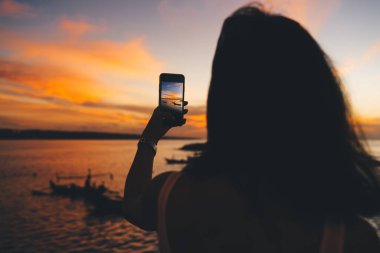 Gün batımında deniz kenarında cep telefonu kullanan kimliği belirsiz kadın turistin arka planı.