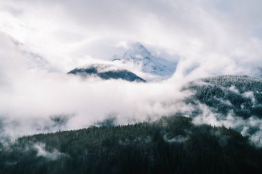 Kışın kasvetli bir gökyüzü altında beyaz buzla kaplı görkemli dağların üzerinde süzülen kalın bulutların nefes kesici drone görüntüsü.