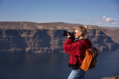 Sırt çantası ve modern fotoğraf makinesiyle gülümseyen mutlu kadın turistin yan görüntüsü. Amerika 'daki muhteşem nehrin yüksek kayalık kıyılarında tatil yaparken fotoğraf çekiyor.