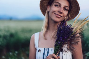 Yeşil çayırda dikilen ve elinde bir demet taze çiçek tutarken gülümseyen sıradan giyinmiş genç bir kadın.