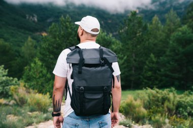 Sırt çantalı, yeşil ormanın yakınında yeşil bir ormanda dikilen, sıradan tişörtlü ve şapkalı yüzsüz bir adam.