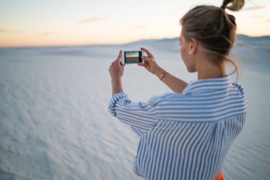 Beyaz Kumlar Ulusal Parkı 'nda gün batımının fotoğrafını çeken akıllı telefonlu bir kadın gezgin çölün ortasında kumlu zeminde duruyor.