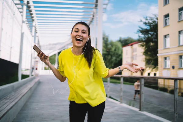 Positive asian female in casual wear laughing on funny content listening audio book on smartphone,smiling woman in headphones holding mobile phone laughing entertaining with music outdoors