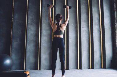 Egzersiz sırasında ağırlık kaldıran kaslı bir kadın figürünün arka planında spor salonundaki aletleri kullanarak kollarını kaldıran bir kadının mükemmel vücut şeklini koruması.