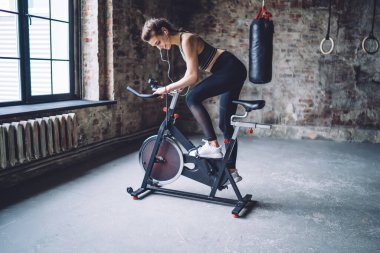 Örgülü saçlı, spor elbiseli bisiklet süren ve kulaklıkla müzik dinleyen genç bir kadının yan görüntüsü.