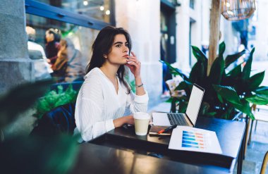 Proje sırasında dizüstü bilgisayarları incelerken resmi kıyafetler içinde, kafenin ahşap masasında oturan düşünceli kadın serbest çalışanın yan görüntüsü