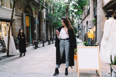 Uzun boylu, kahverengi saçlı, şık ceketli, cep telefonlu, SMS 'e cevap veren ve şehir caddesinde yürürken boş afişin yanında duran genç bir kadın.