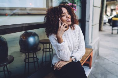 Kol saati takılı gülümseyen Afro-Amerikalı kadın ahşap bankta oturuyor ve sokakta dinlenirken akıllı telefondan konuşuyor.