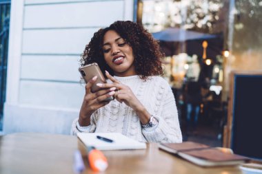 Gülümseyen koyu tenli kıvırcık hippi kız akıllı telefondan video izlediği için 4G bağlantısının keyfini çıkarıyor, Afro-Amerikan milenyum kadını cep telefonuyla web sayfalarında kitap yazıyor.