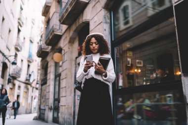 Şehir caddesinde elinde not defteri ve cep telefonuyla duran şık kıyafetli, odaklanmış genç kız öğrencilerden.