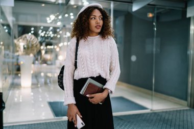 Şık giyimli ve bereli Afro-Amerikan bayan akıllı telefon ve defterle modern alışveriş merkezinde duruyor ve başka tarafa bakıyor.