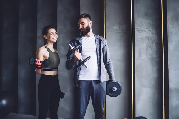 积极的年轻女性穿着运动服 与男朋友一起在健身房锻炼力量 微笑的女性对职业胡子训练员的工作感到满意 表现出她们能举起哑铃 — 图库照片