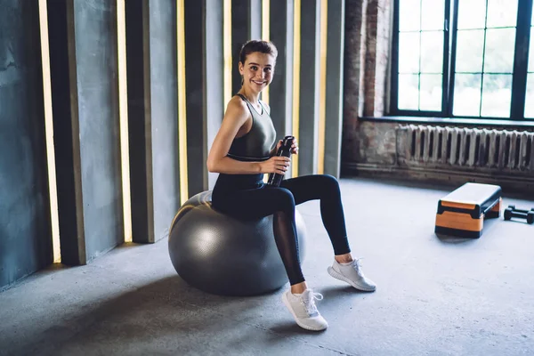 Full body of slim smiling female in sportswear sitting on fit ball with bottle of water and looking at camera in gym