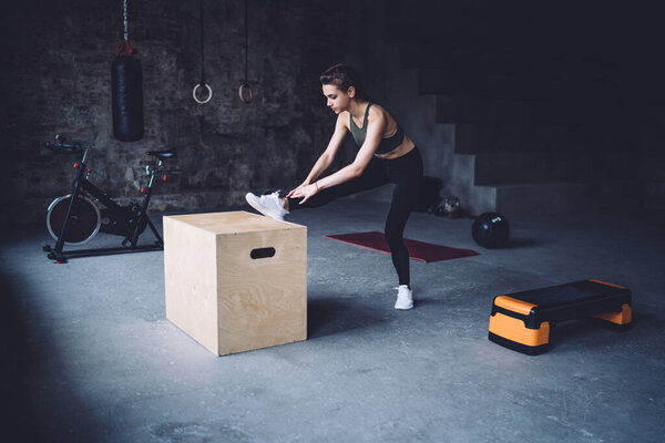 Full length side view of fit young woman putting leg on wooden box for stretching tense muscles after step aerobics training