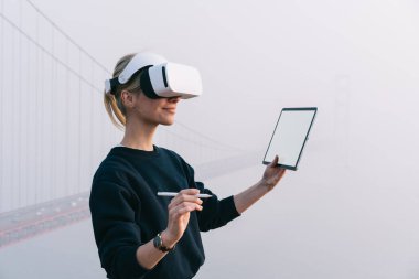 İş sırasında elinde boş tablet ve sanal kalemle tepede duran, at kuyruklu, sanal kulaklı bayanın yan görüntüsü.