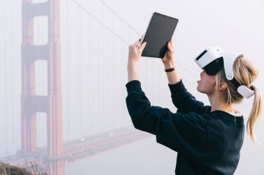 San Fransisco 'da köprünün dışında VR gözlük ve tablet kullanan sıradan giysili sarışın kadının yan görüntüsü.