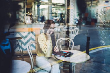 Moda gözlüklü ve parlak sarı kazaklı genç ve çekici bir kadının samimi modern kafede planlamacıyla otururken arkadaşını aramasının yan görüntüsü.
