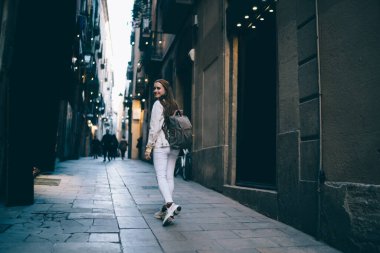 Sırt çantalı genç bayan Barcelona 'nın yaşlı caddesinde kaldırımda yürürken sırtüstü kameraya bakıp gülümsüyor.