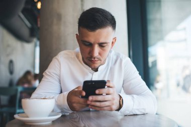 Kafede bir fincan kahveyle ahşap masada otururken beyaz gömlekli ciddi bir erkek cep telefonuyla sörf yapıyor.