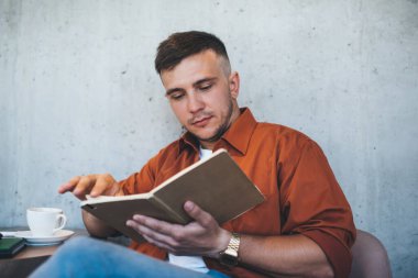 Modern kafede bir fincan kahveyle bulanık cephede dinlenirken rahat rahat giyinen adam ilginç bir kitap okuyor.