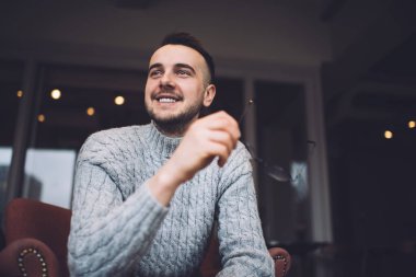 Sıradan giyinmiş neşeli sakallı bir adam, gözlük takıyor ve rahat bir kafede dinlenip gülümserken başka tarafa bakıyor.