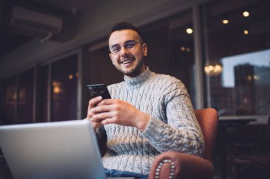 Düşük açılı, sakallı, serbest çalışan, sıradan kıyafetler ve gözlüklü, gülümseyen ve akıllı telefondan sörf yaparken kameraya bakan bir erkek.