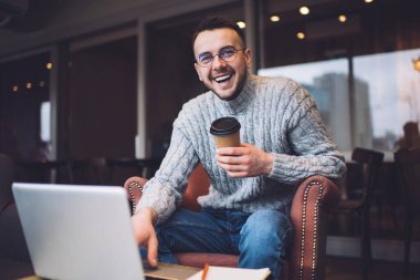 Sıcak süveter giyen, rahat koltukta oturan ve molada bir bardak sıcak içecek içerken bilgisayar kullanan mutlu bir erkek serbest yazar.