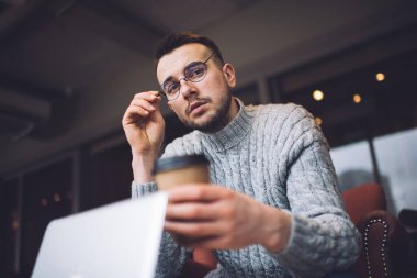 Ciddi bir genç adam gözlükleriyle yuvarlak ayarlar ve kahve içerken kameraya bakar.