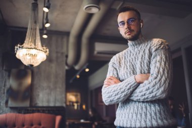 Gri, sıcak kazaklı ve kablosuz kulaklıklı genç adam kollarını kavuşturup restoranda dinlenirken kameraya bakıyor.