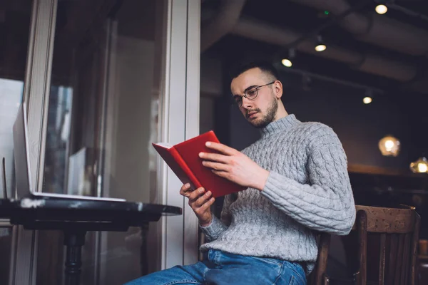 穿着保暖衣服和眼镜的严肃大胡子男子坐在木椅上 一边在现代咖啡店的笔记本电脑上读书 一边在写日记 — 图库照片