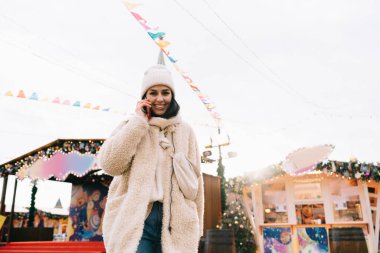 Sıcak giyimli, şapkalı, gülümseyen ve cep telefonuyla konuşurken kameraya bakan iyimser bir kadından.