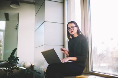 Gözlüklü neşeli bayan öğrencinin pencerenin kenarında oturup netbook ve cep telefonuyla sınavlara hazırlanmasından.