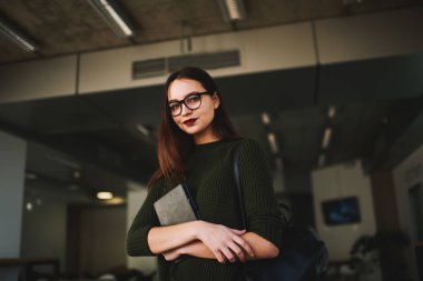 Gözlüklü, gözlüklü, sırt çantalı, rahat bir odada kameraya bakarken elinde not defteri olan neşeli bir kadın.