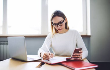 Konsantre genç işçi masada oturuyor ve bilgisayar ve telefon kullanırken not defterine notlar yazıyor ve aydınlık odada gözlüklü sıradan kıyafetler giyiyor.