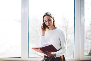 Gündüz vakti pencere eşiğinde oturan, telefonla konuşan ve not defterinden notlar okuyan ciddi bir uzaktan kumanda işçisi.