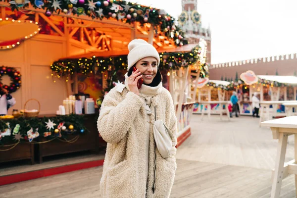 ガーランドやライトで飾られた街の中心部を歩いている間にスマートフォンで会話をしながら暖かいコートで陽気な若い女性観光客 — ストック写真